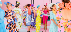 Women dressed in vibrant, stylish outfits at the Melbourne Cup, featuring bold floral dresses, colorful fascinators, and statement hats against a pink and white checkered backdrop.